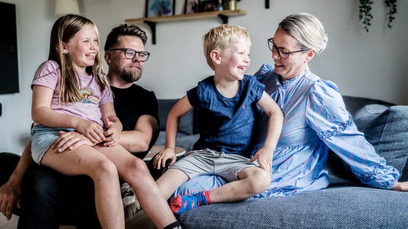 Billedet viser en familie bestående af to voksne og to børn. De to børn sidder på skødet af deres far og mor. Hele familien griner