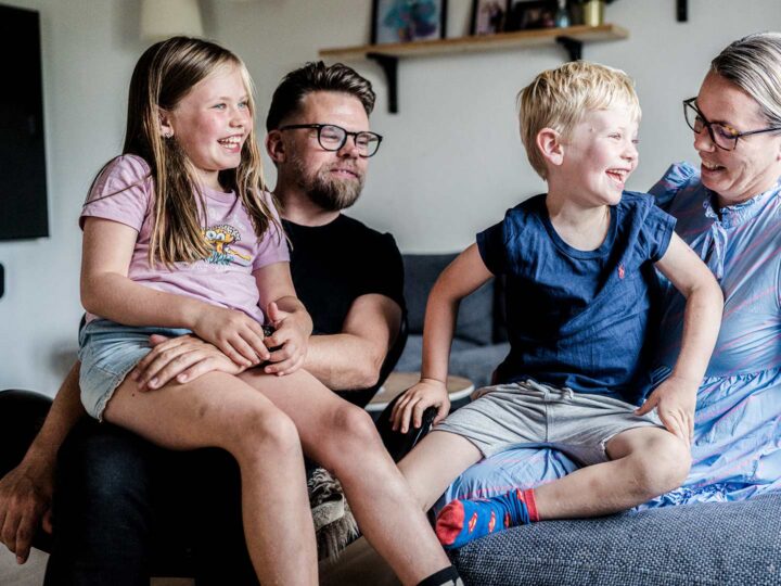 Billedet viser en familie bestående af to voksne og to børn. De to børn sidder på skødet af deres far og mor. Hele familien griner